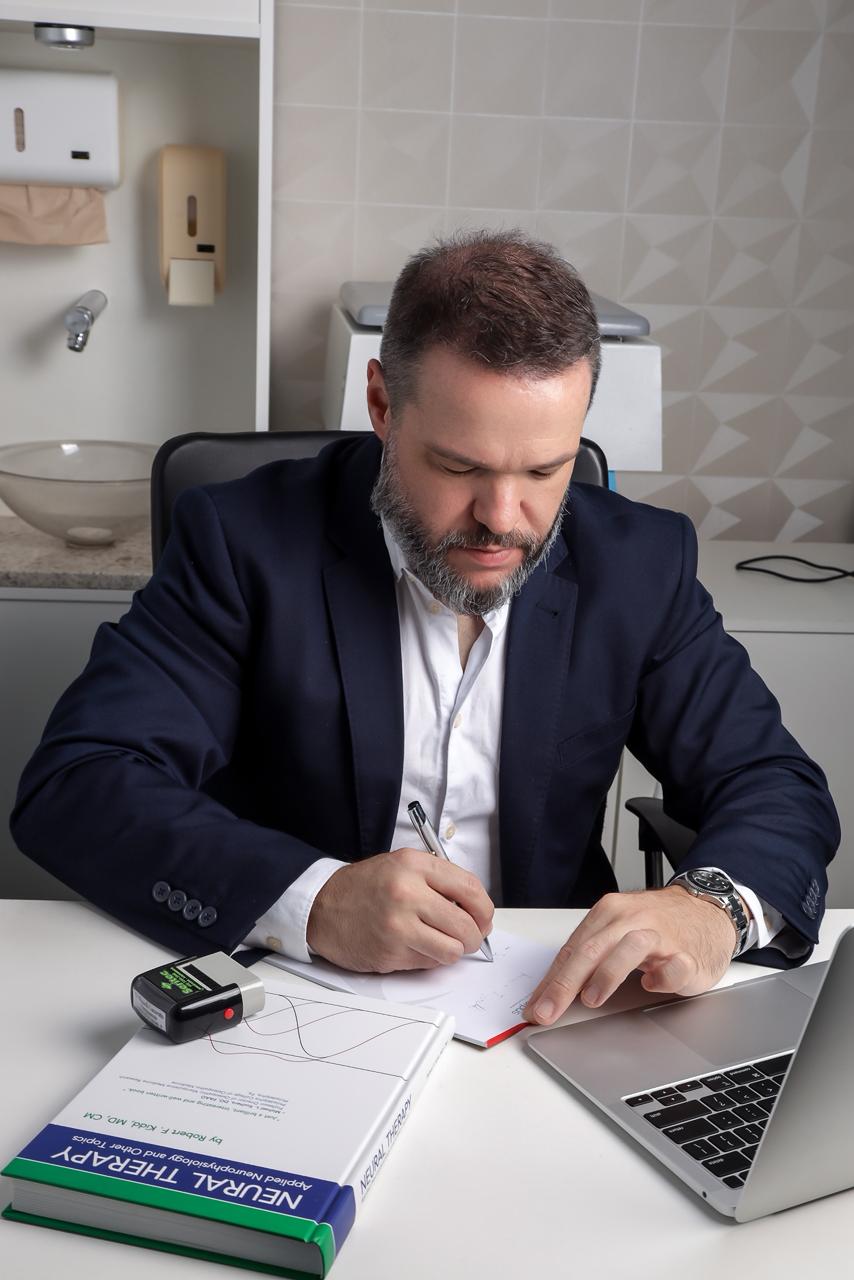 Dr. Ricardo concentrado, escrevendo à mão sobre a mesa do consultório