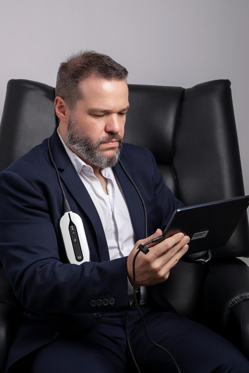 Dr. Ricardo sentado numa poltrona de couro, consultando um tablet conectado a um aparelho de ultrassom portátil